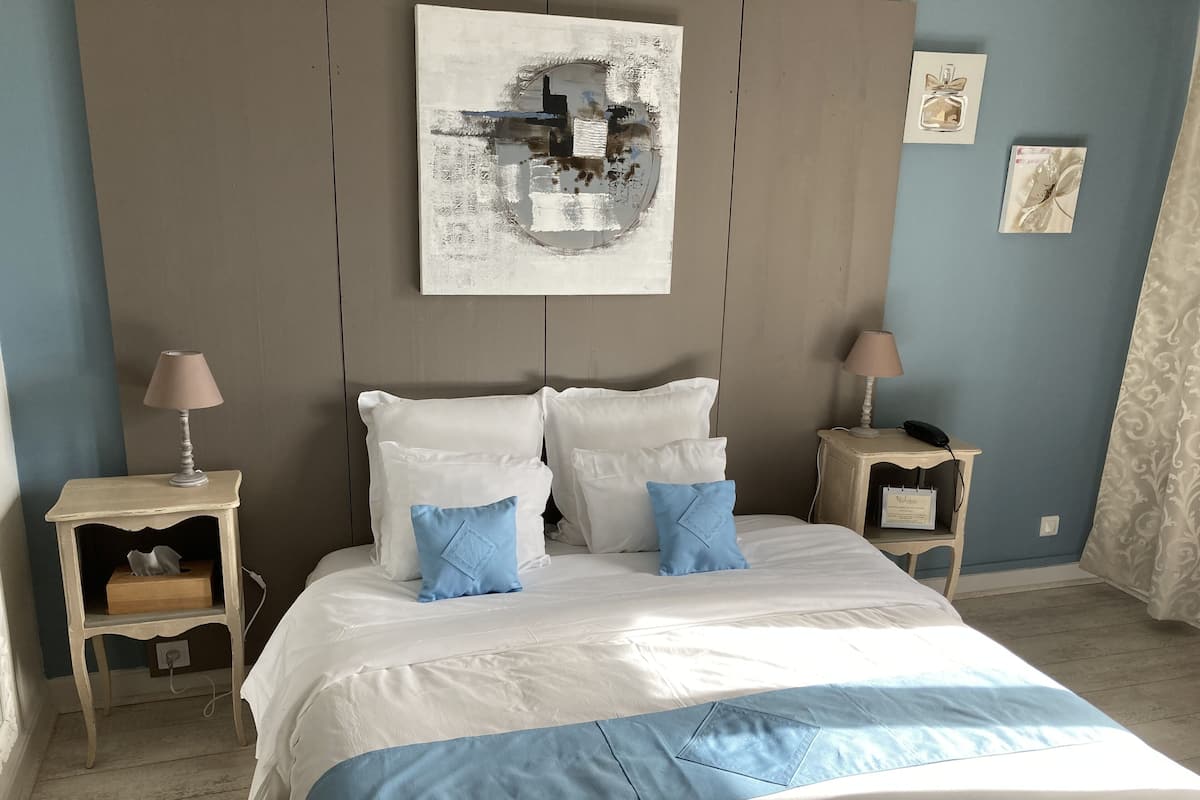 Une chambre à coucher bien aménagée, avec un grand lit, deux tables de chevet, une œuvre d’art accrochée au mur et un rideau à motifs.
