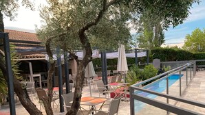 An outdoor patio area with tables and chairs, a swimming pool, and olive trees. - Logis Hôtel les Aubuns Country Hôtel (Saint-Gilles)