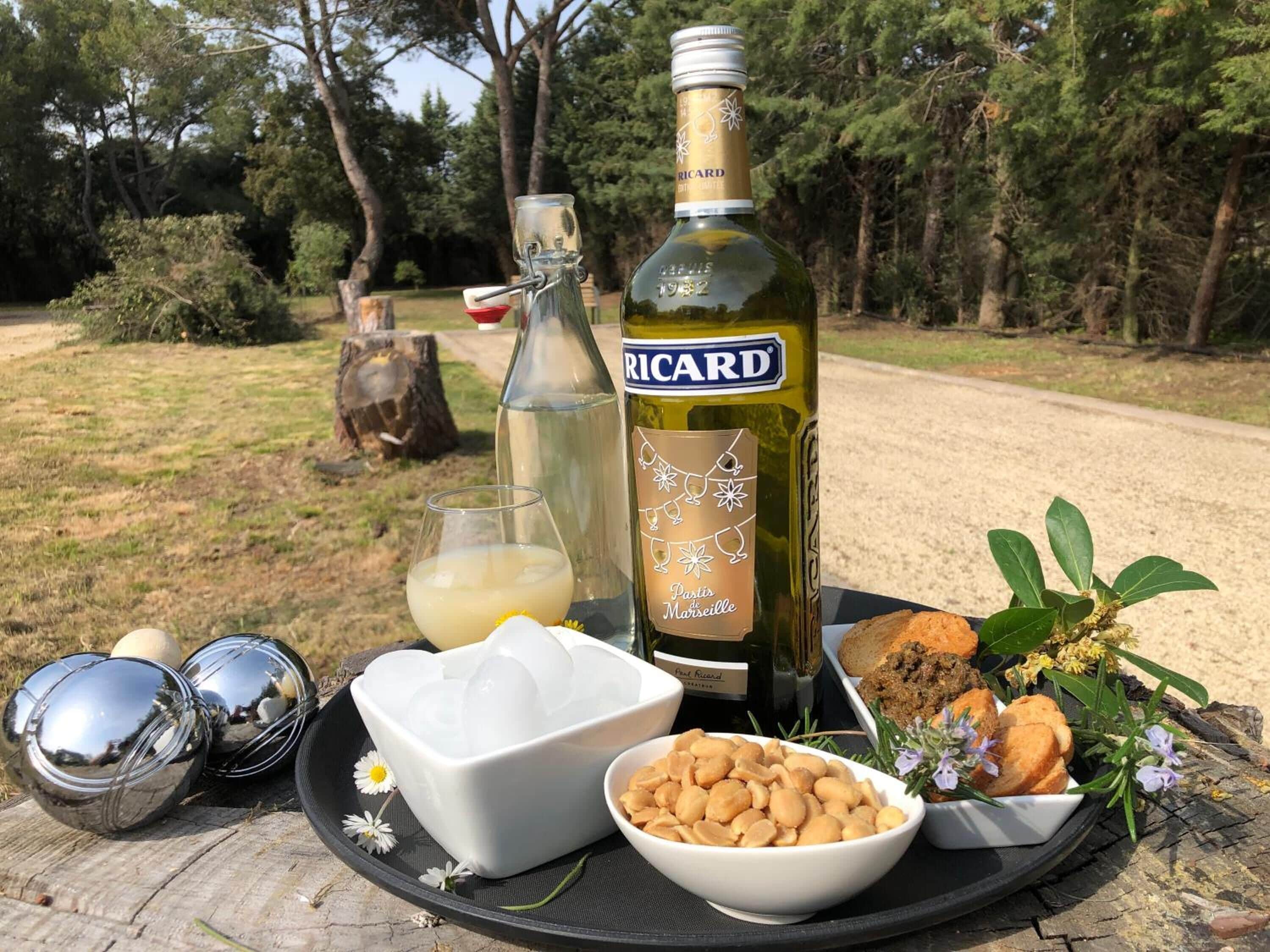 A picnic setup with a bottle of Ricard wine, a glass of white wine, a bowl of nuts, a plate with cookies, and decorative flowers on a wooden table outdoors.