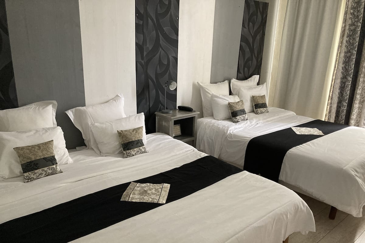 Une chambre d’hôtel avec deux lits, chacun agrémenté de draps blancs et de coussins décoratifs. Une table de chevet avec une lampe se trouve entre les deux lits.