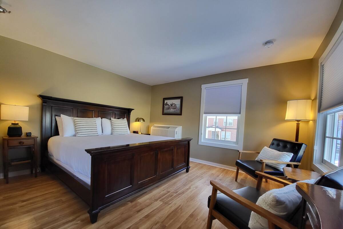 A hotel room with a large bed, bedside lamps, a chair, a window with blinds, and a radiator.