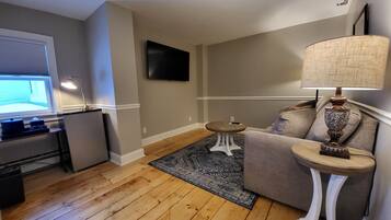 A living room with a wooden floor, a grey sofa, a small white side table with a lamp, and a flat-screen TV mounted on the wall.