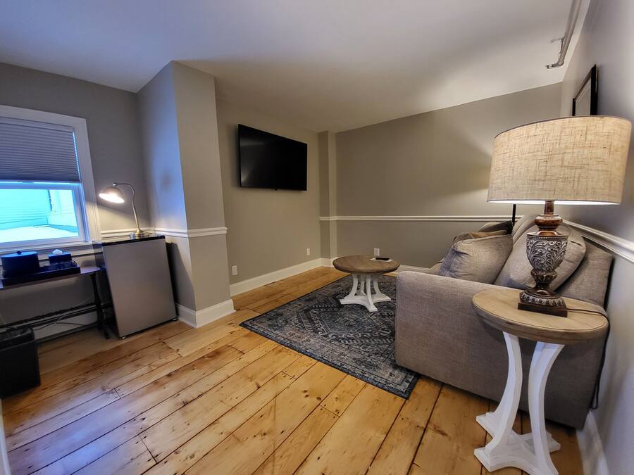 A living room with a wooden floor, a grey sofa, a small white side table with a lamp, and a flat-screen TV mounted on the wall.
