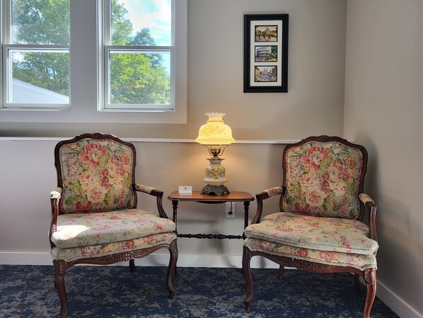 Two ornate chairs with floral upholstery, a table with a lamp, and a framed picture on the wall.