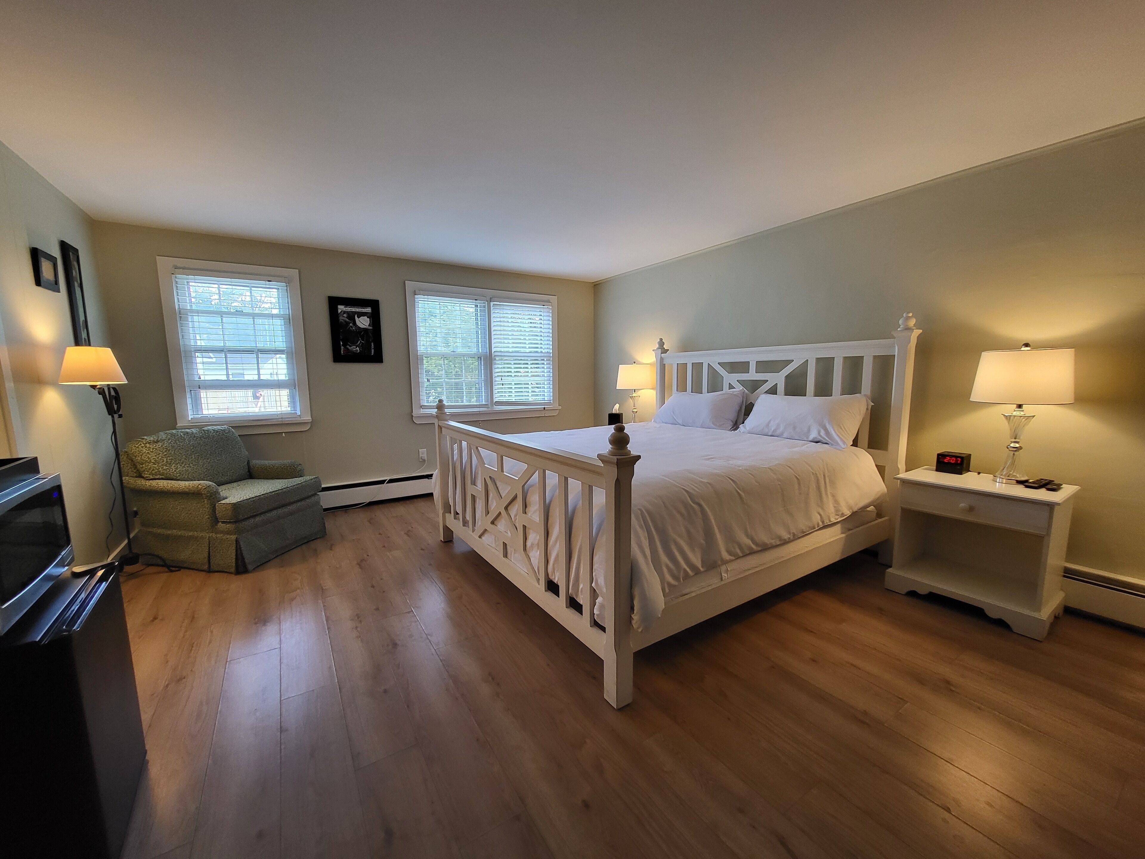 Un dormitorio con una cama blanca, dos mesitas de noche con lámparas, una silla y un televisor.