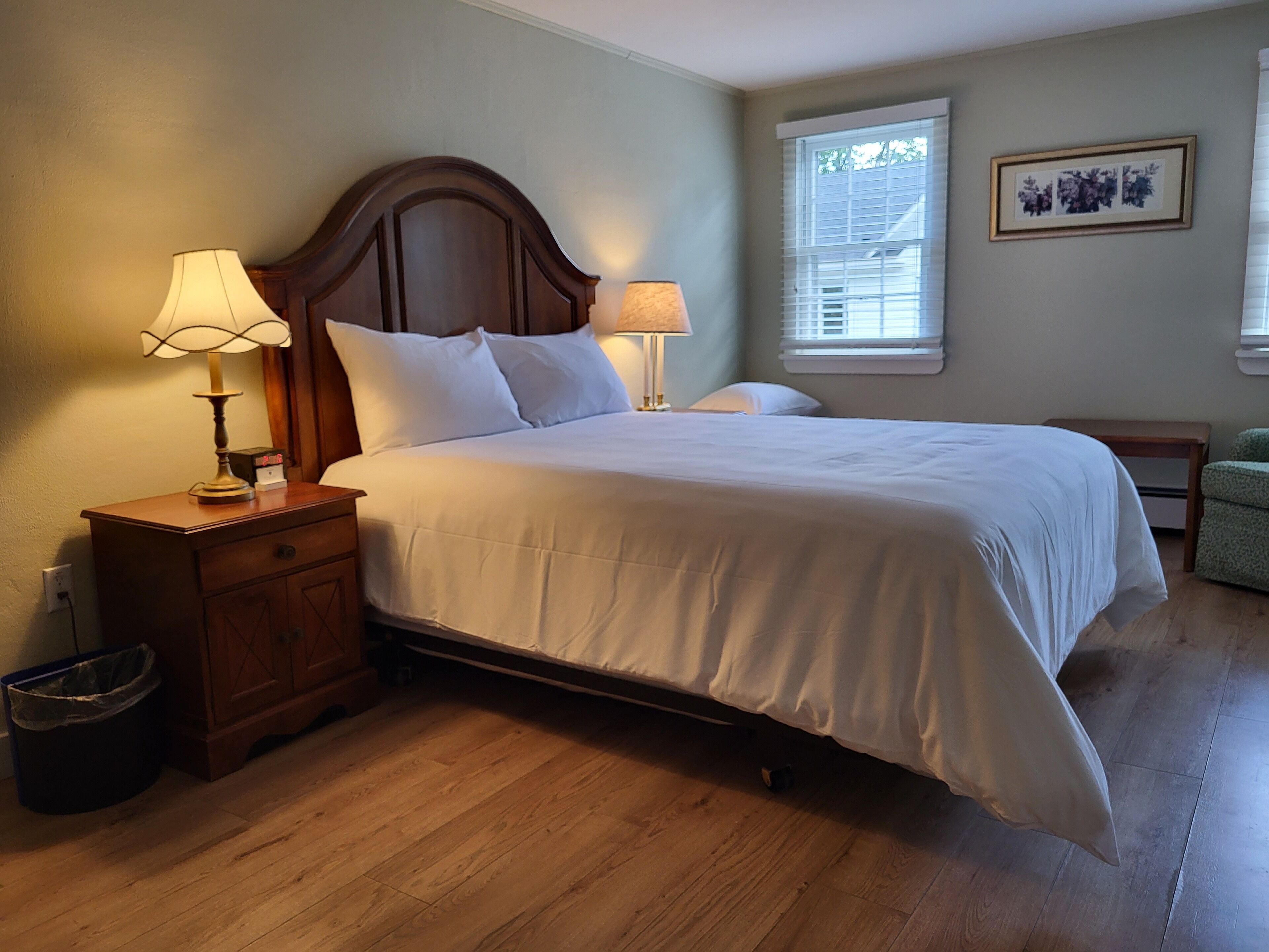A neatly made bed in a room with wooden flooring, two bedside tables with lamps, a trash bin, and a window with blinds.
