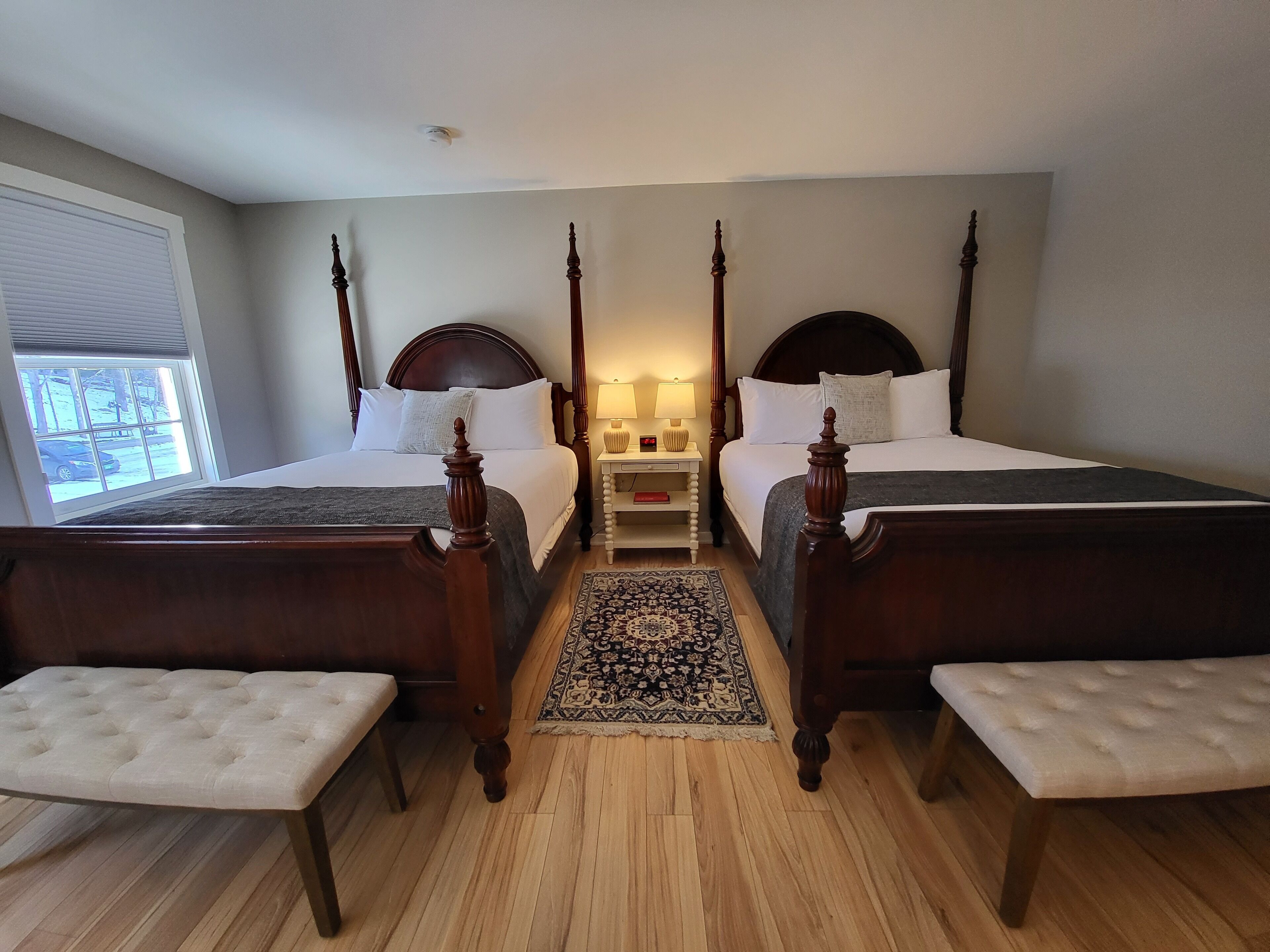 A hotel room with two beds, a nightstand, a bedside table, a window with blinds, and a view of the outside.