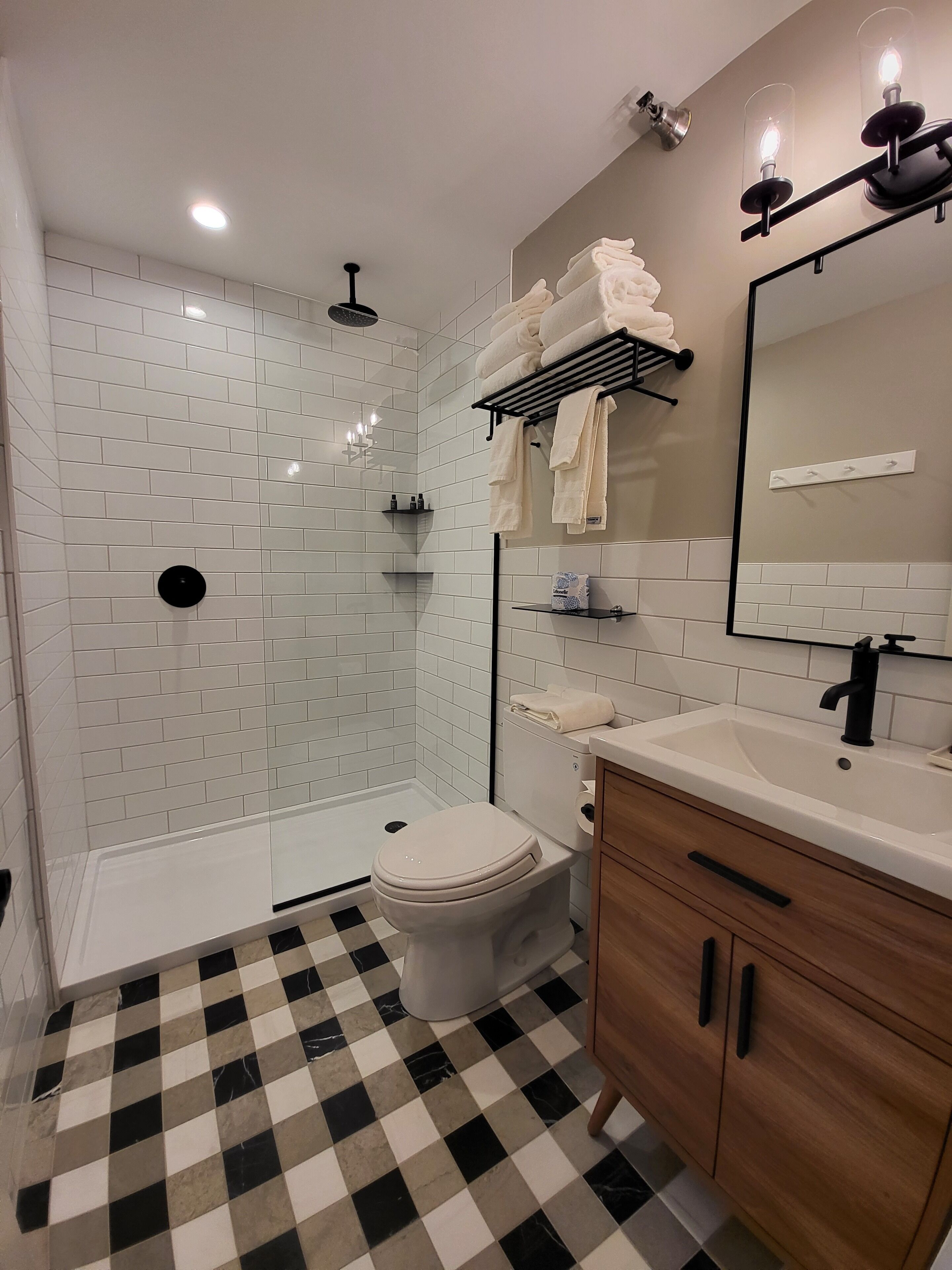 Un baño moderno con azulejos blancos tipo metro, piso a cuadros blanco y negro, inodoro blanco, tocador de madera con espejo y zona de ducha con puerta de vidrio.