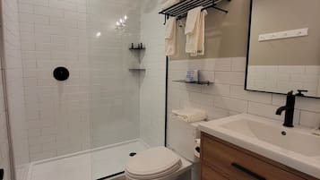 Un baño moderno con azulejos blancos tipo metro, piso a cuadros blanco y negro, inodoro blanco, tocador de madera con espejo y zona de ducha con puerta de vidrio.