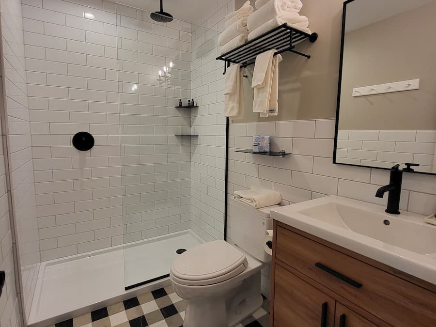 A modern bathroom with white subway tiles, a black and white checkered floor, a white toilet, a wooden vanity with a mirror, and a shower area with a glass door.
