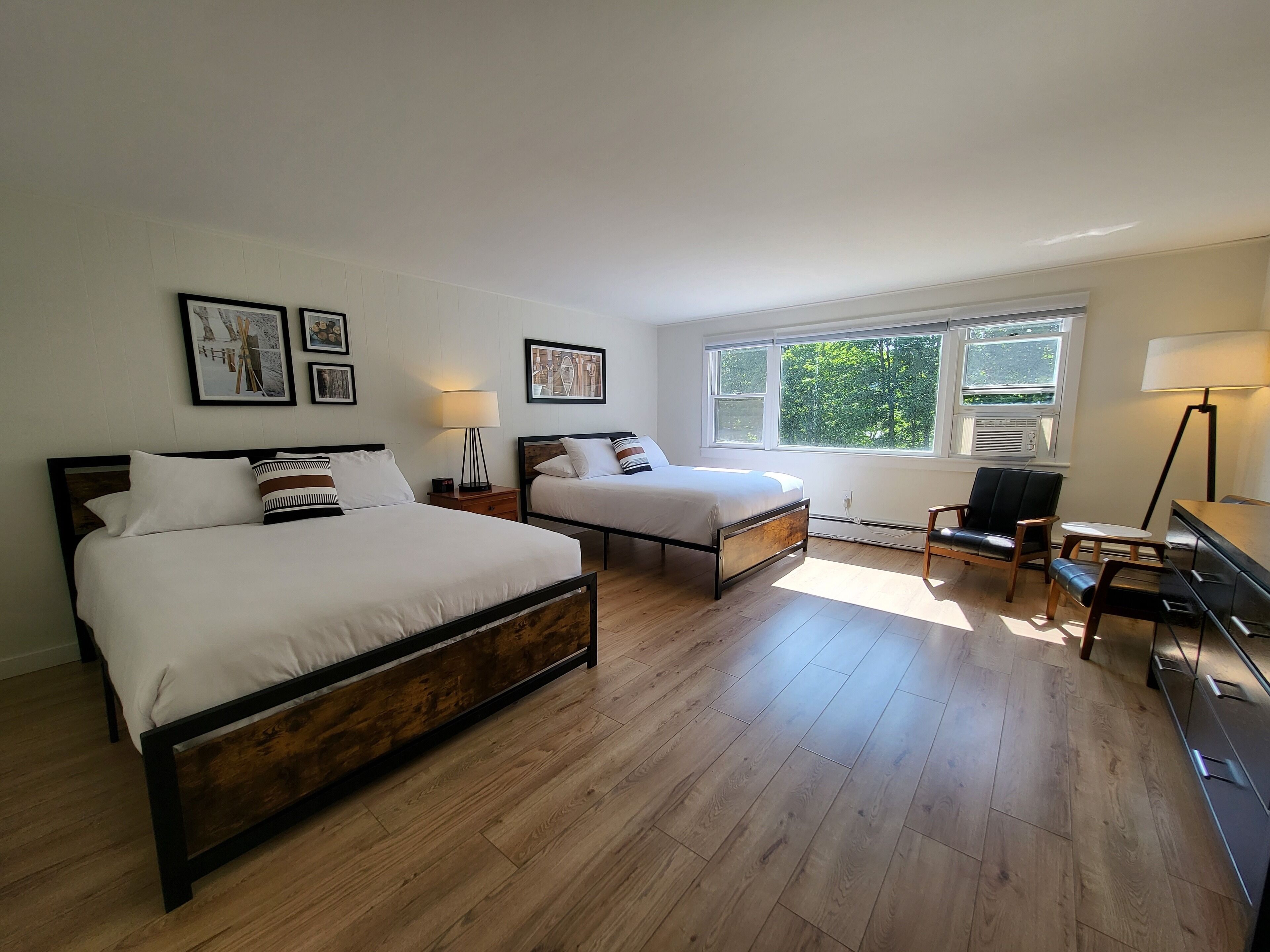 A bedroom with two beds, a desk, a chair, and a window with a view of greenery.