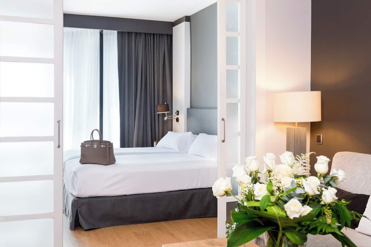 A hotel room with a bed, a bedside table with a lamp, a vase of flowers, and a view of the outside.