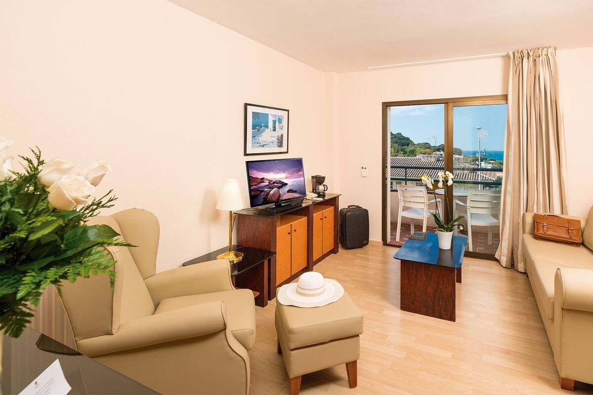 Een hotelkamer met een bank, fauteuil, salontafel en een bureau met een computer. Er is een raam met uitzicht op zee en een balkon met stoelen.