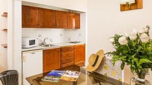 Une cuisine avec des meubles de rangement en bois, un four à micro-ondes et un évier. On y trouve une table avec des magazines et un chapeau. Un vase avec des fleurs blanches est posé sur la droite.