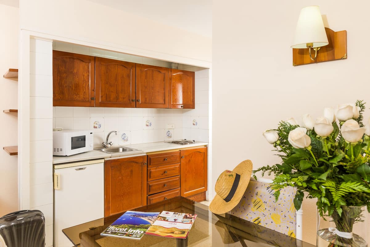 Een keuken met houten kasten, een magnetron en een spoelbak. Er staat een tafel met tijdschriften en een hoed. Een vaas met witte bloemen staat aan de rechterkant.