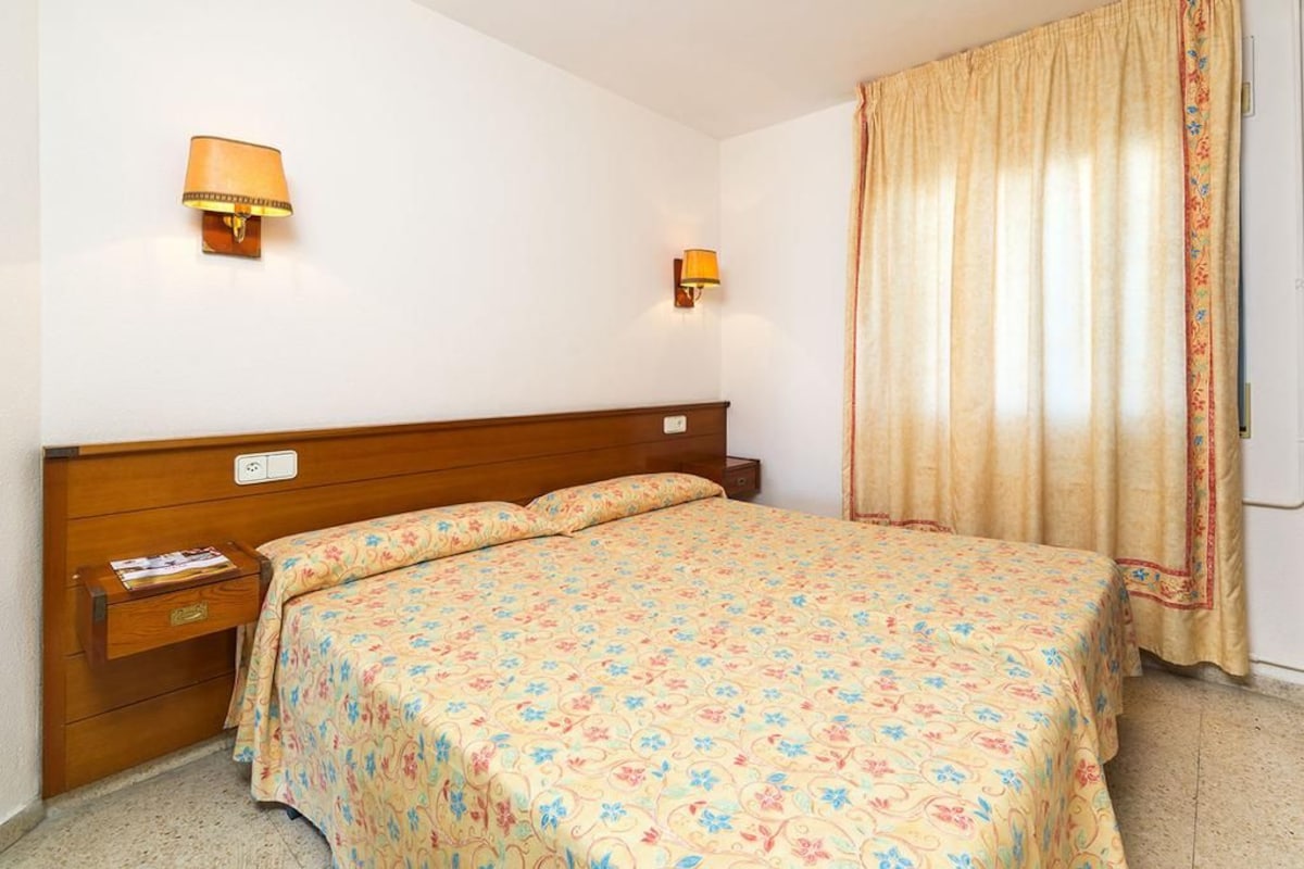 Habitación de hotel con cabecera de madera, dos lámparas de noche, colcha estampada y ventana con cortinas.