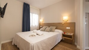 Un lit bien fait, avec des oreillers et une serviette pliée, une table de chevet avec une lampe et un magazine, et une fenêtre avec des rideaux.