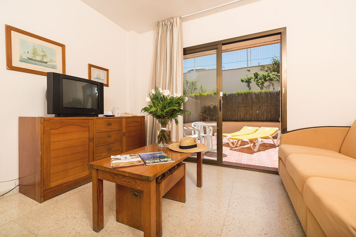 Een kamer met een houten kast, een televisie, een tafel met tijdschriften, een hoed en een plant. Er is een schuifdeur van glas die leidt naar een buitenruimte met gele stoelen en een tafel.