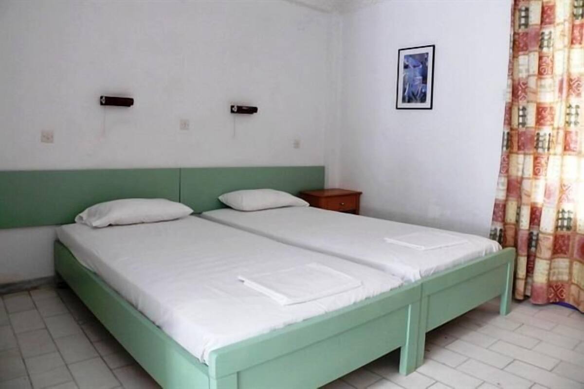 A room with two single beds, a small wooden nightstand, a framed picture on the wall, and a patterned curtain.