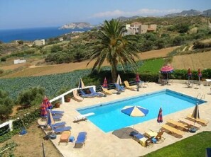 A pool area with lounge chairs and umbrellas, surrounded by greenery and a view of the sea and hills. - Esperides Apartments (Malevizi)
