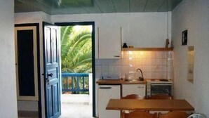 A compact kitchen with a dining area, a view of a palm tree through an open door, and a small sink. - Esperides Apartments (Malevizi)