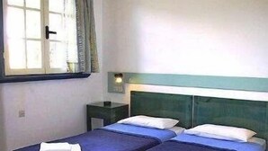 A room with two single beds, each with a blue bedspread and white pillows, a nightstand with a lamp, and a window with curtains. - Esperides Apartments (Malevizi)