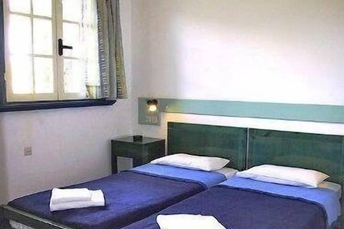 A room with two single beds, each with a blue bedspread and white pillows, a nightstand with a lamp, and a window with curtains.