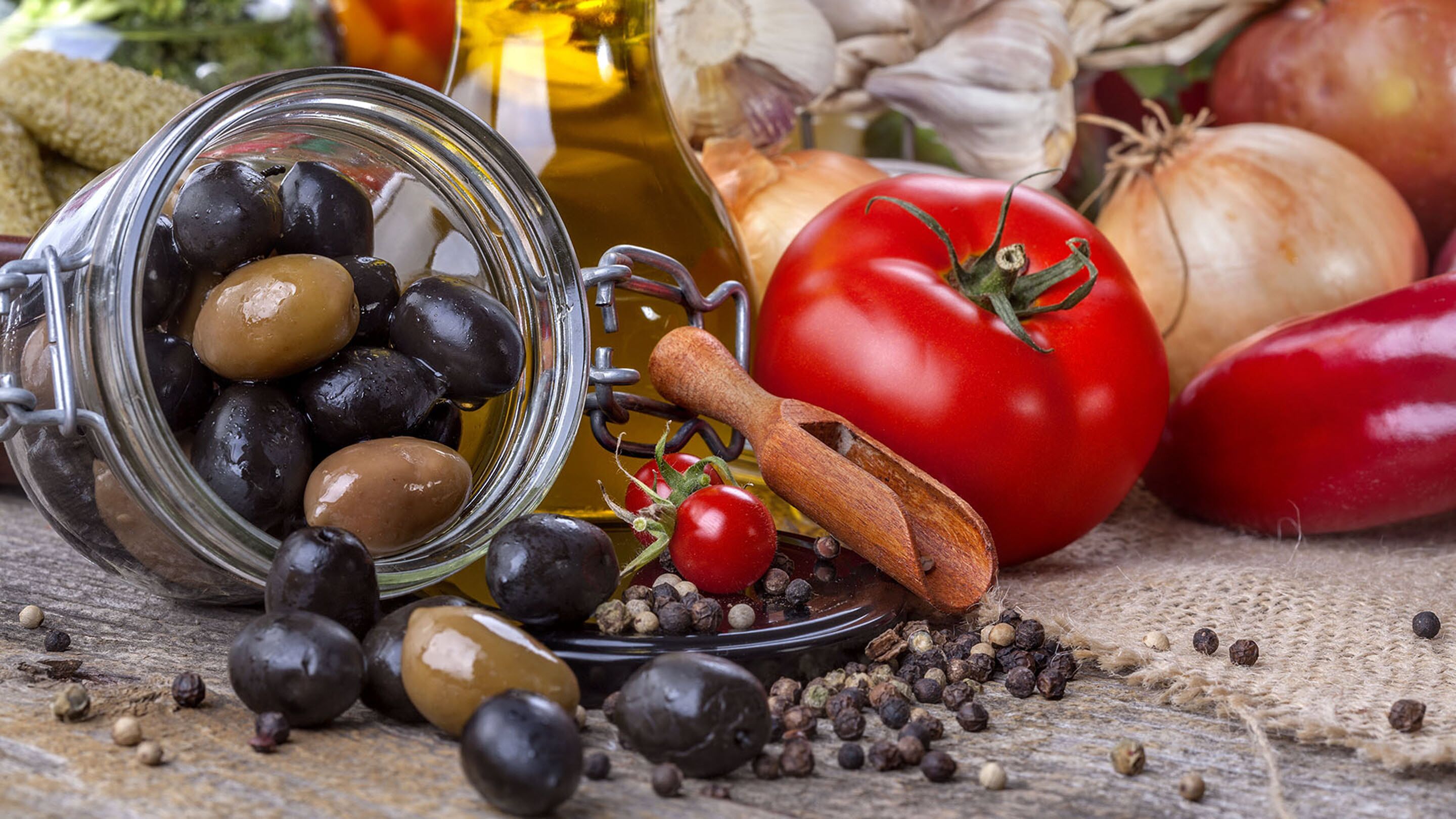 A jar of olives, a tomato, a pepper mill, and onions on a rustic surface.