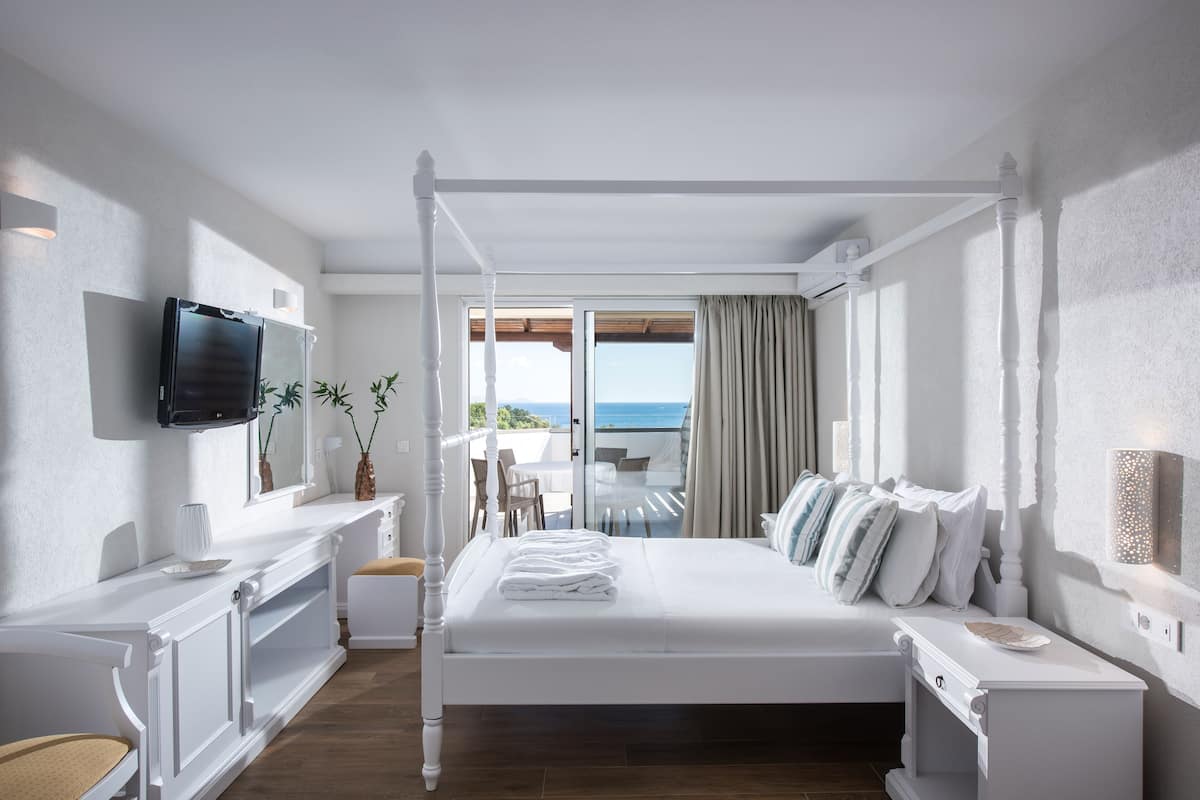 A bedroom with a four-poster bed, a television, a desk, and a view of the ocean.