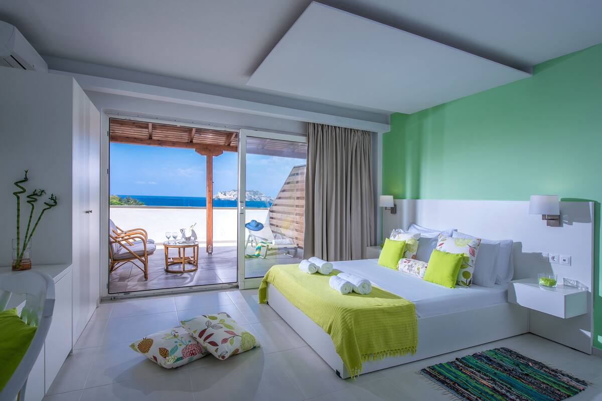 A modern hotel room with a large bed, a balcony with a view of the sea, and a separate seating area with a table and chairs.