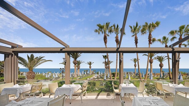 A terrace with tables and chairs, overlooking a beach with palm trees and umbrellas. - Creta Star Hotel (Rethymno)