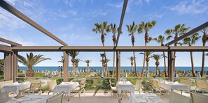 Una terraza con mesas y sillas, con vista a una playa con palmeras y sombrillas.