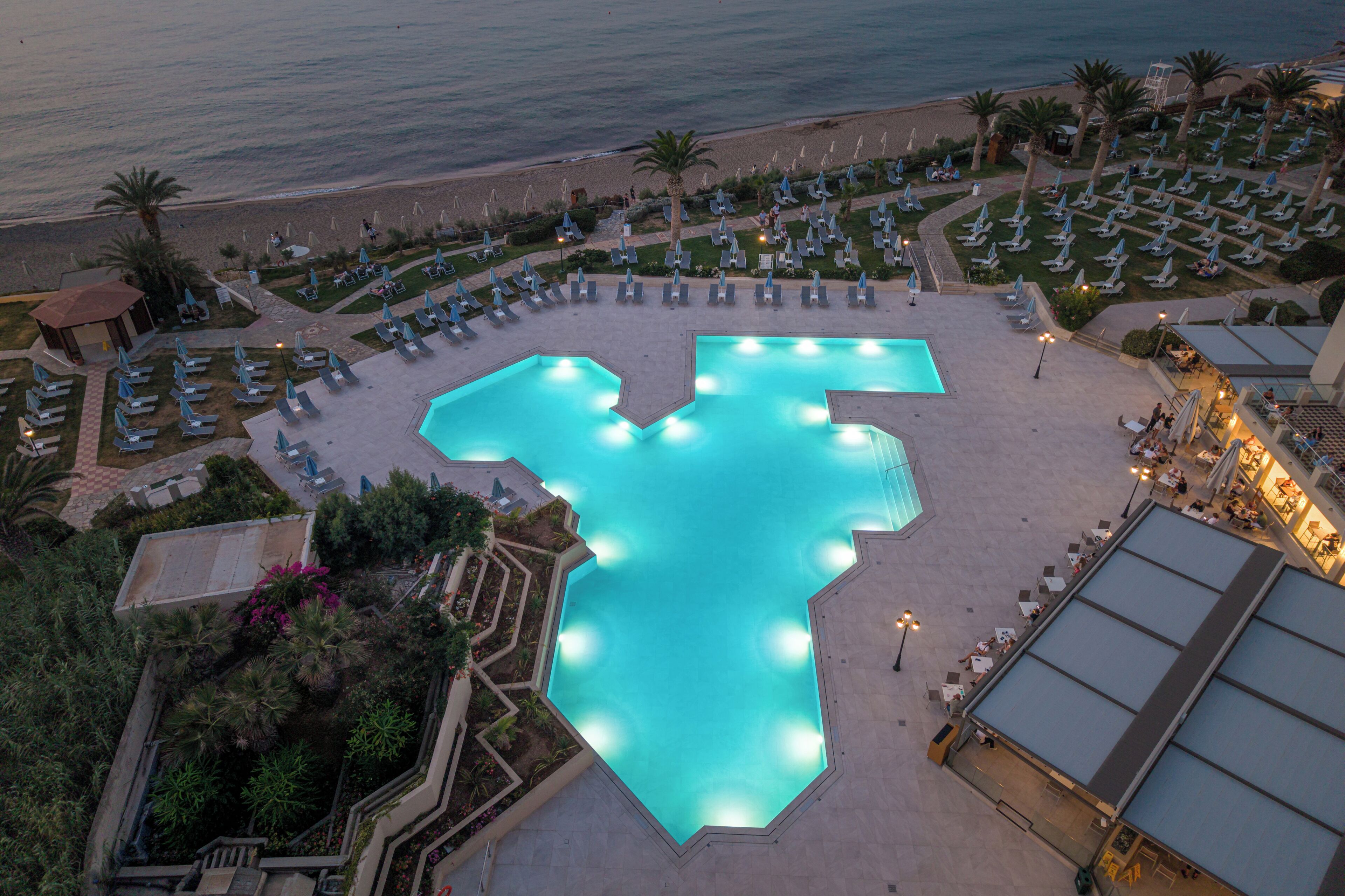 Vista aérea de un complejo turístico con una gran piscina en forma de letra 'M', rodeada de tumbonas y una zona de playa.