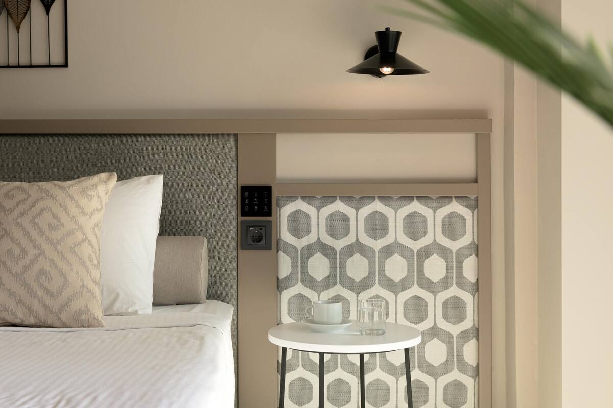 A hotel room with a bed, a bedside table with a cup and glass, a wall-mounted light, and a patterned curtain.