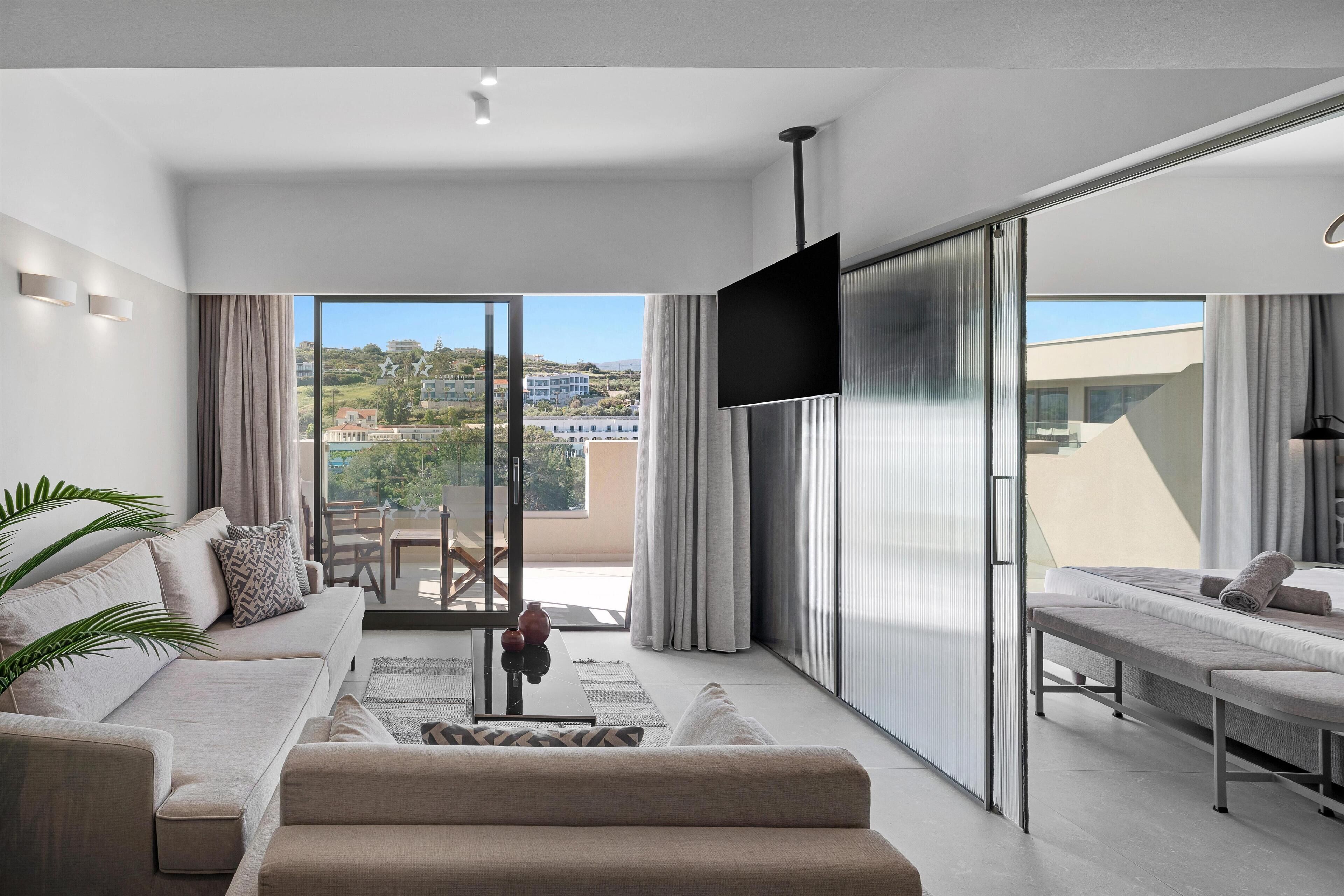 A modern living room with a sofa, a coffee table, and a TV. There is a sliding glass door leading to a balcony with a view of buildings.