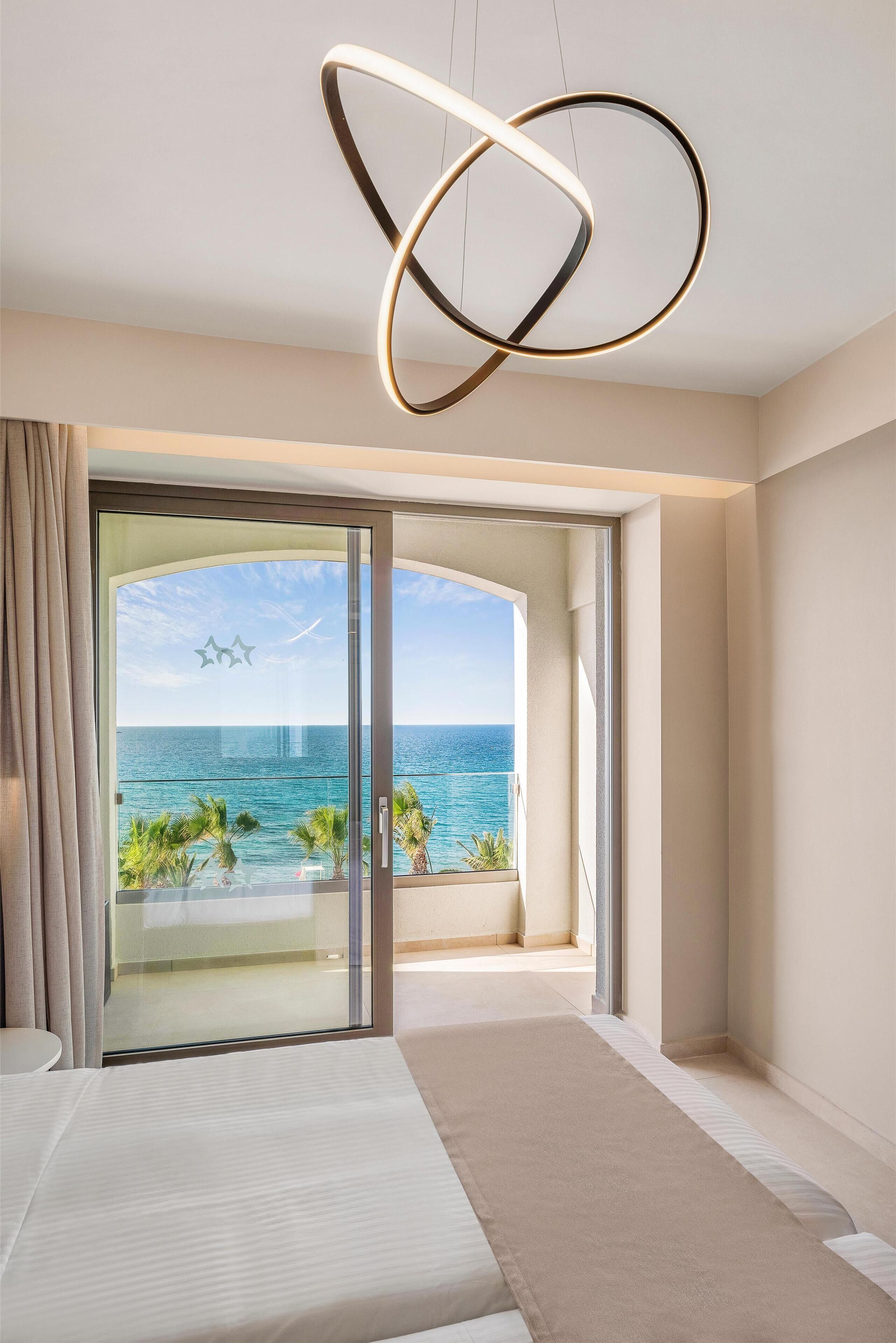 A modern hotel room with a large sliding glass door leading to a balcony with a sea view.