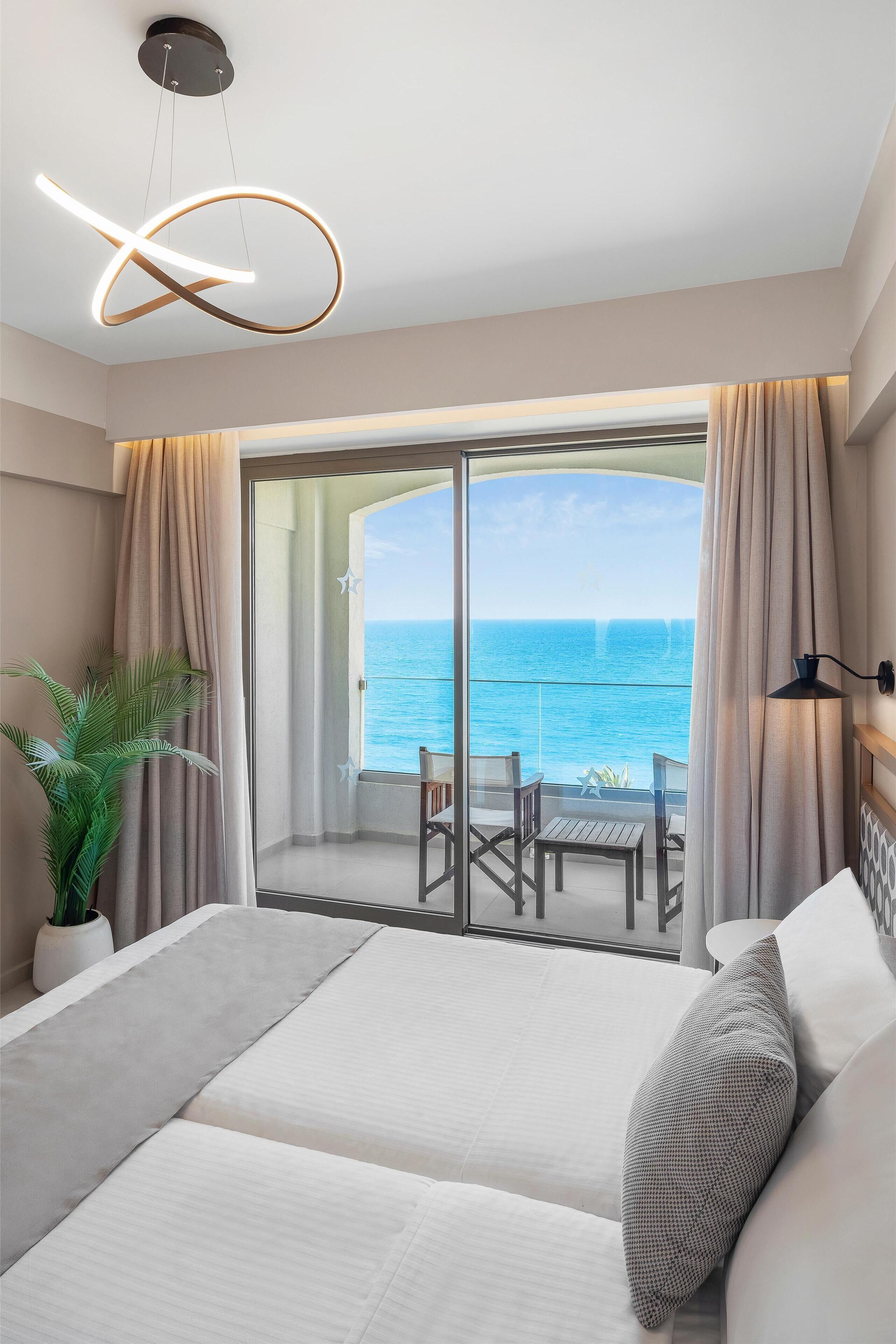 A modern hotel room with a balcony overlooking the sea, featuring a bed, a potted plant, and a view of the ocean.