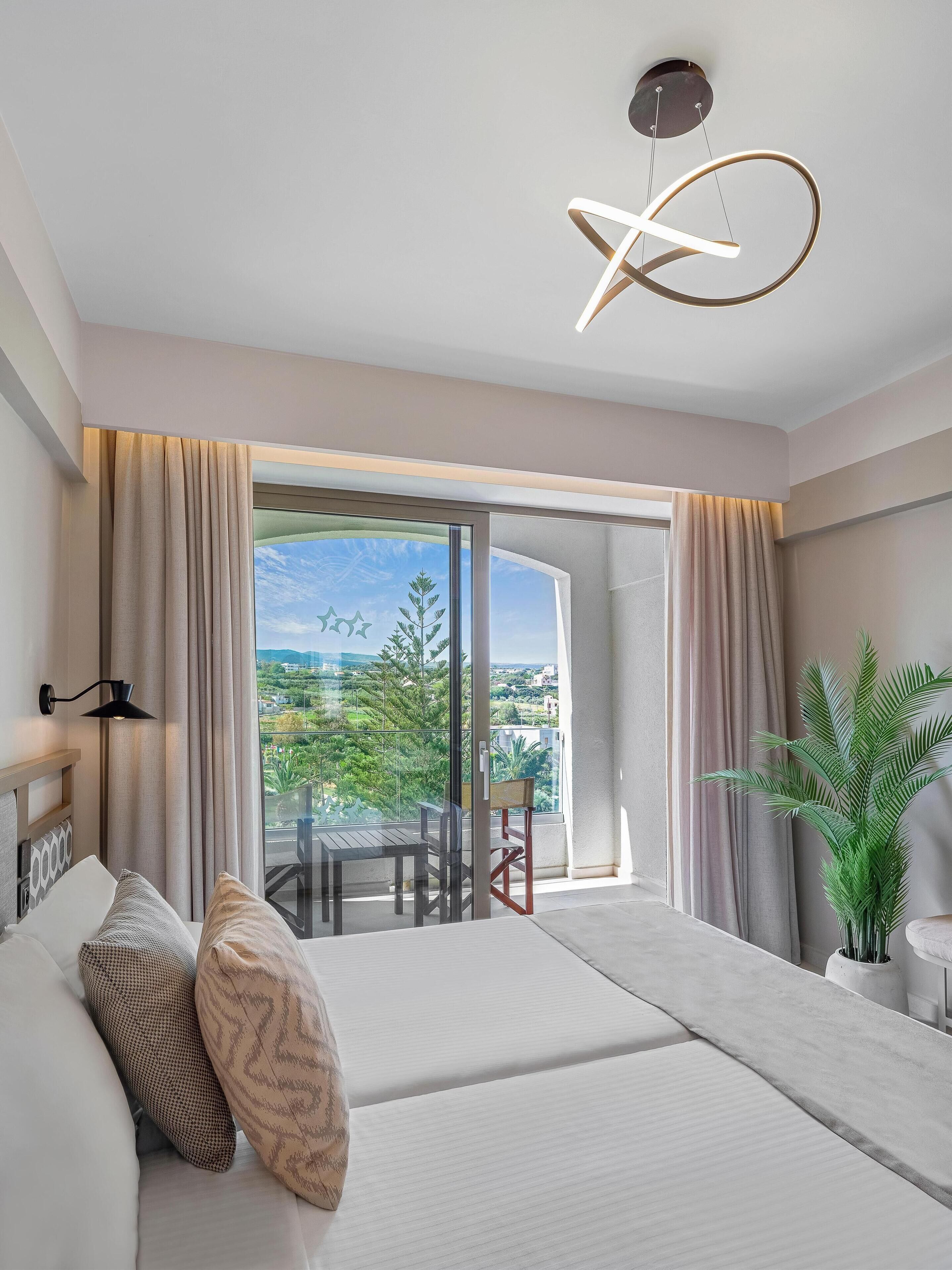 A modern hotel room with a large bed, a balcony with a view of greenery and buildings, a hanging light fixture, and a potted plant.