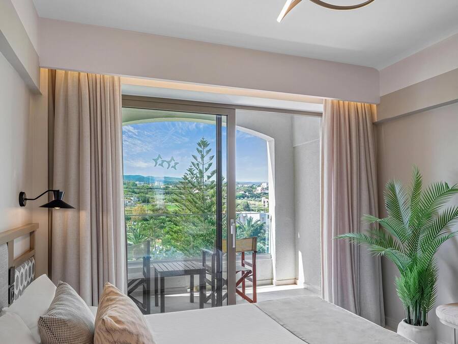A modern hotel room with a large bed, a balcony with a view of greenery and buildings, a hanging light fixture, and a potted plant.