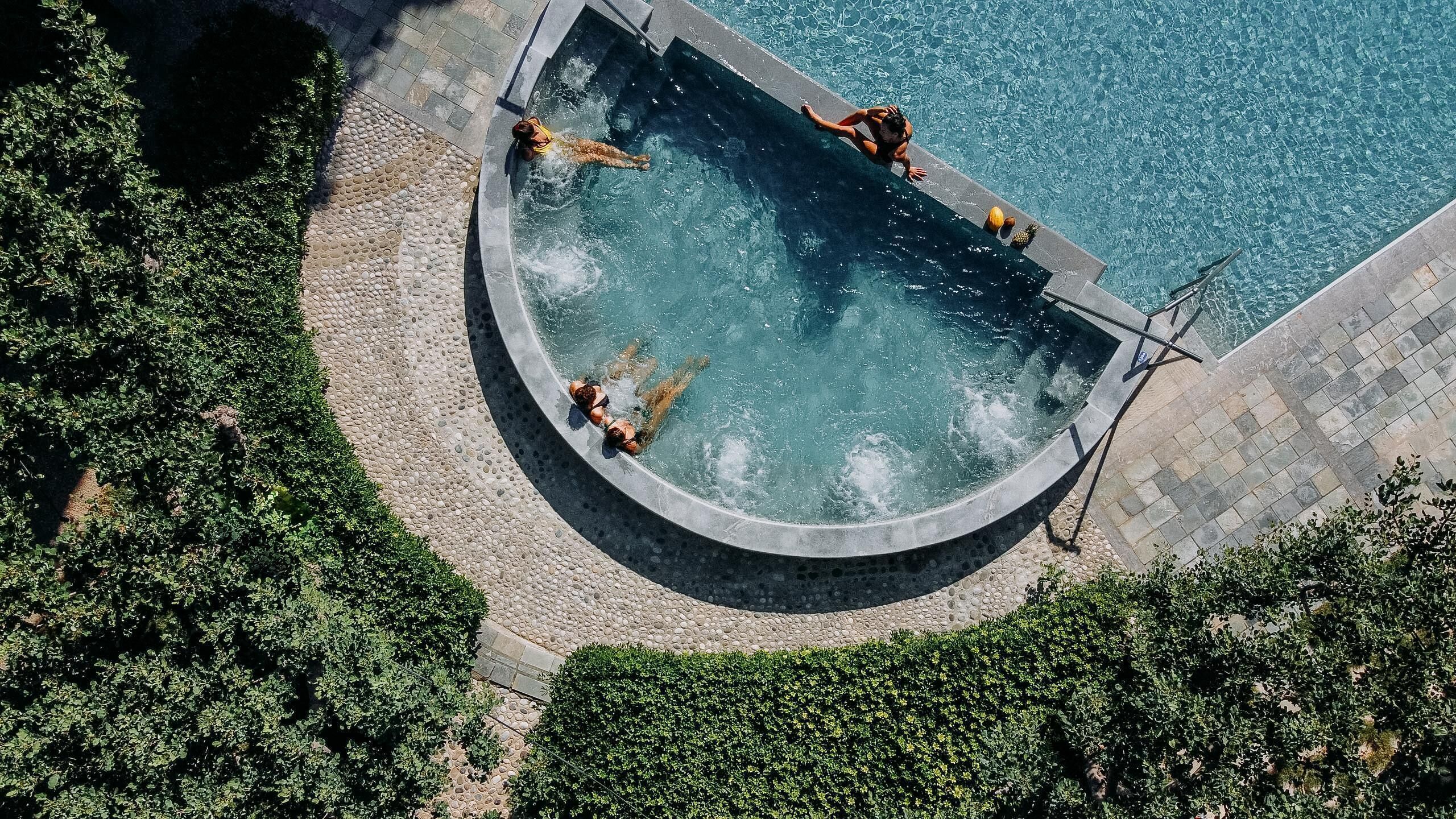 An aerial view of a swimming pool with a curved edge, surrounded by a pebble-lined deck and lush greenery.