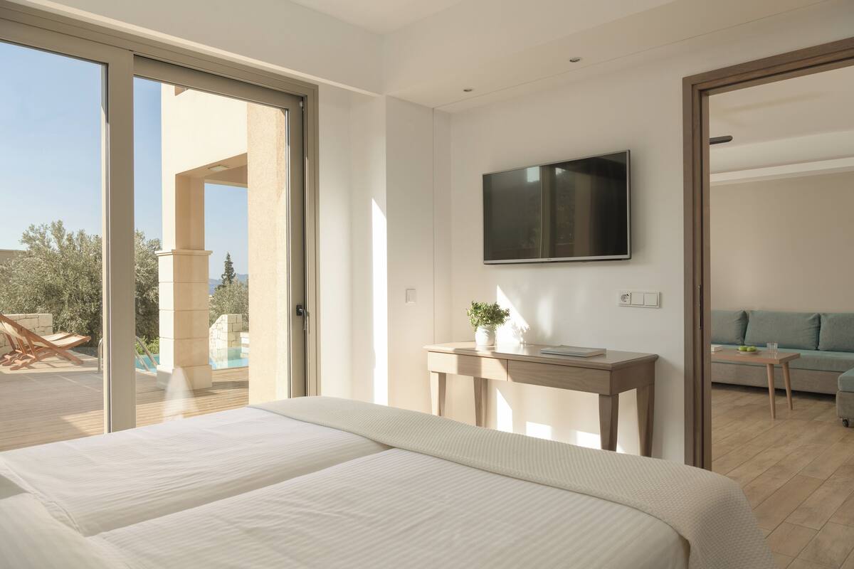 A modern hotel room with a large bed, a TV mounted on the wall, a desk with a plant, and a view of the outdoor pool area.