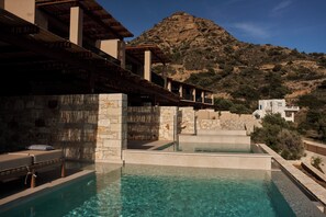 A pool area with a stone wall, a wooden deck, and a mountain in the background. - Paralos Irini Mare (Agios Vasileios)