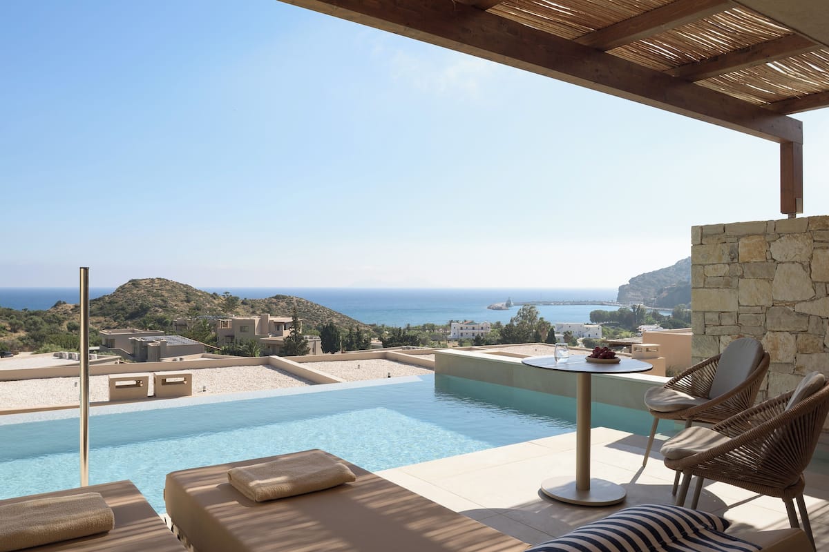A poolside area with lounge chairs, a table, and a view of the sea and hills.