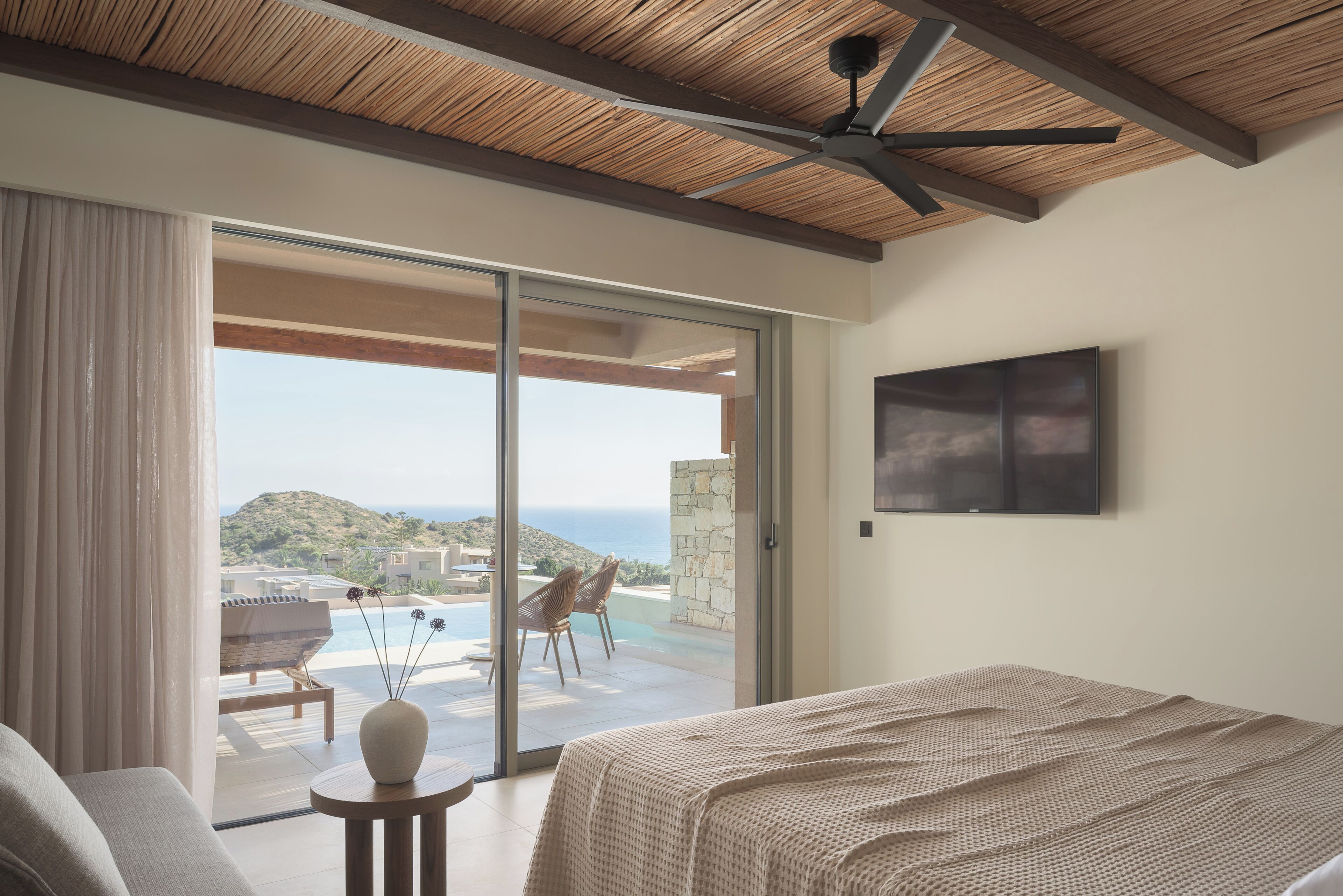 A bedroom with a bed, a ceiling fan, a large window with a view of the sea, and a flat-screen TV on the wall.