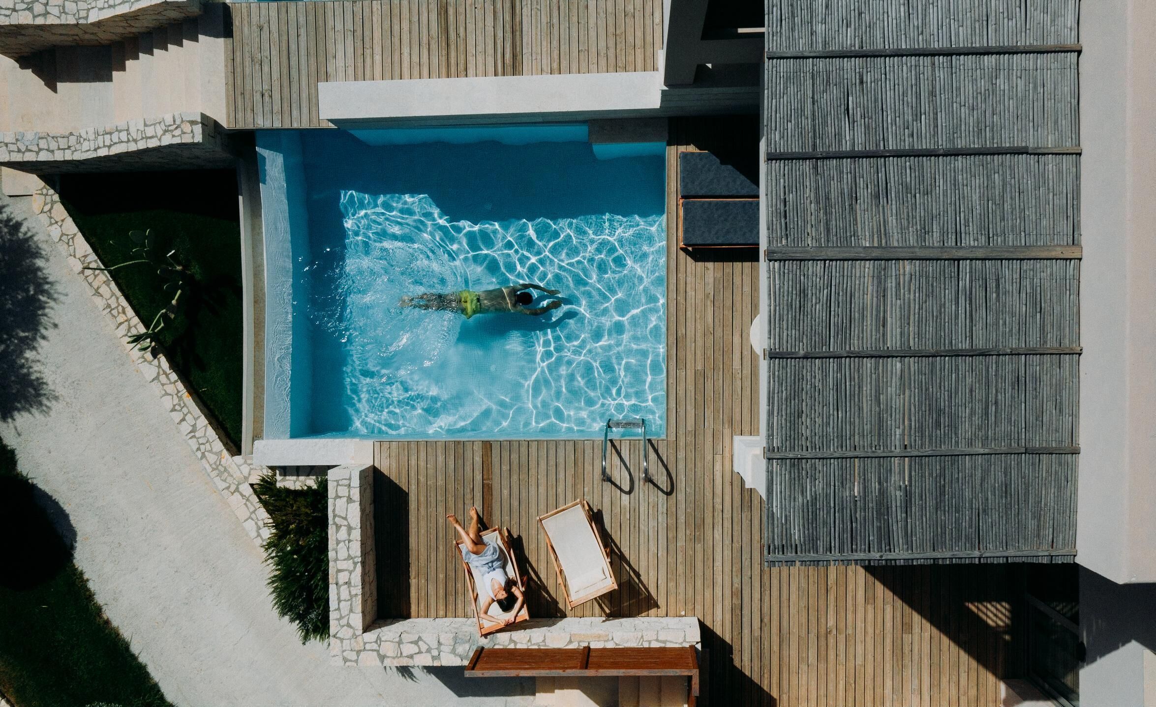 Vista aérea de uma piscina com deck de madeira, área de estar com cadeiras e um espaço coberto para sentar.