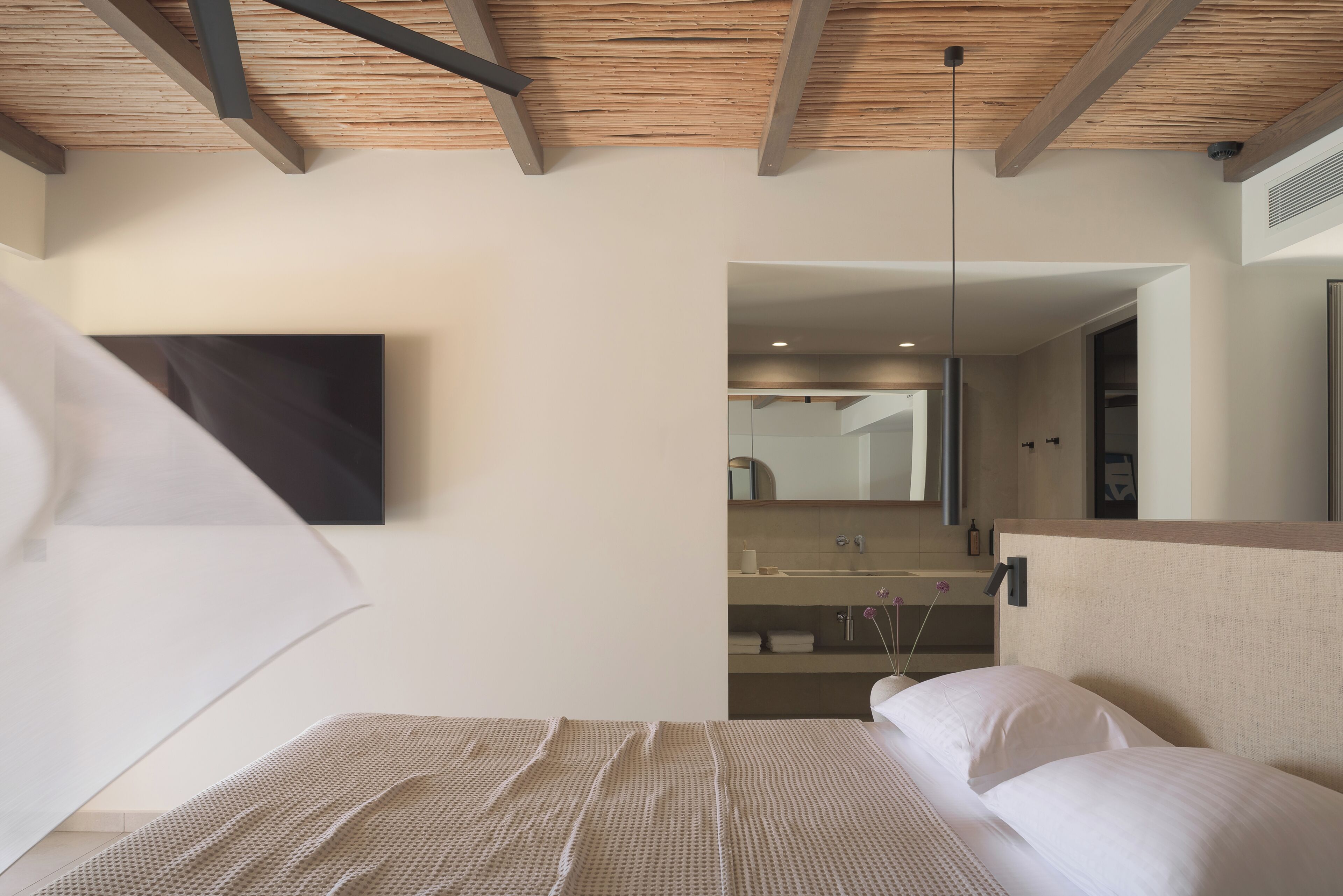 A bedroom with a wooden ceiling, a bed with white bedding, a large mirror, and a bathroom visible through an open door.