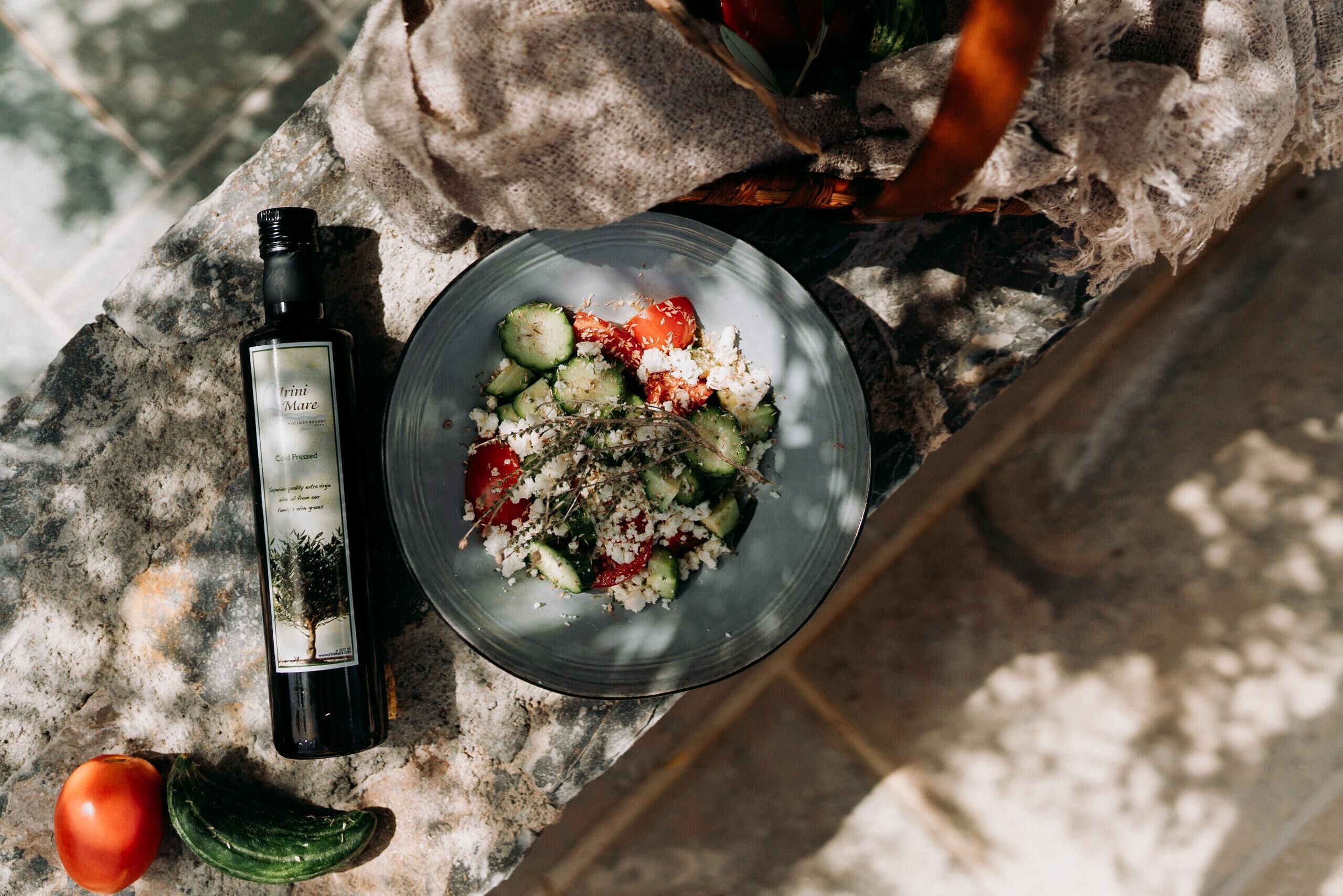 A bottle of wine, a glass bowl with a salad, and a tomato on a stone surface.
