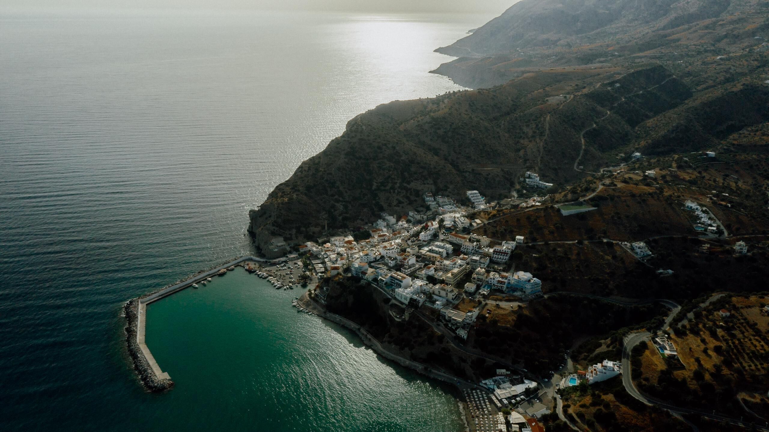 An aerial view of a coastal town with a marina, buildings clustered along the waterfront, and hilly terrain in the background.
