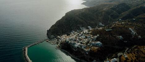 An aerial view of a coastal town with a marina, buildings clustered along the waterfront, and hilly terrain in the background.