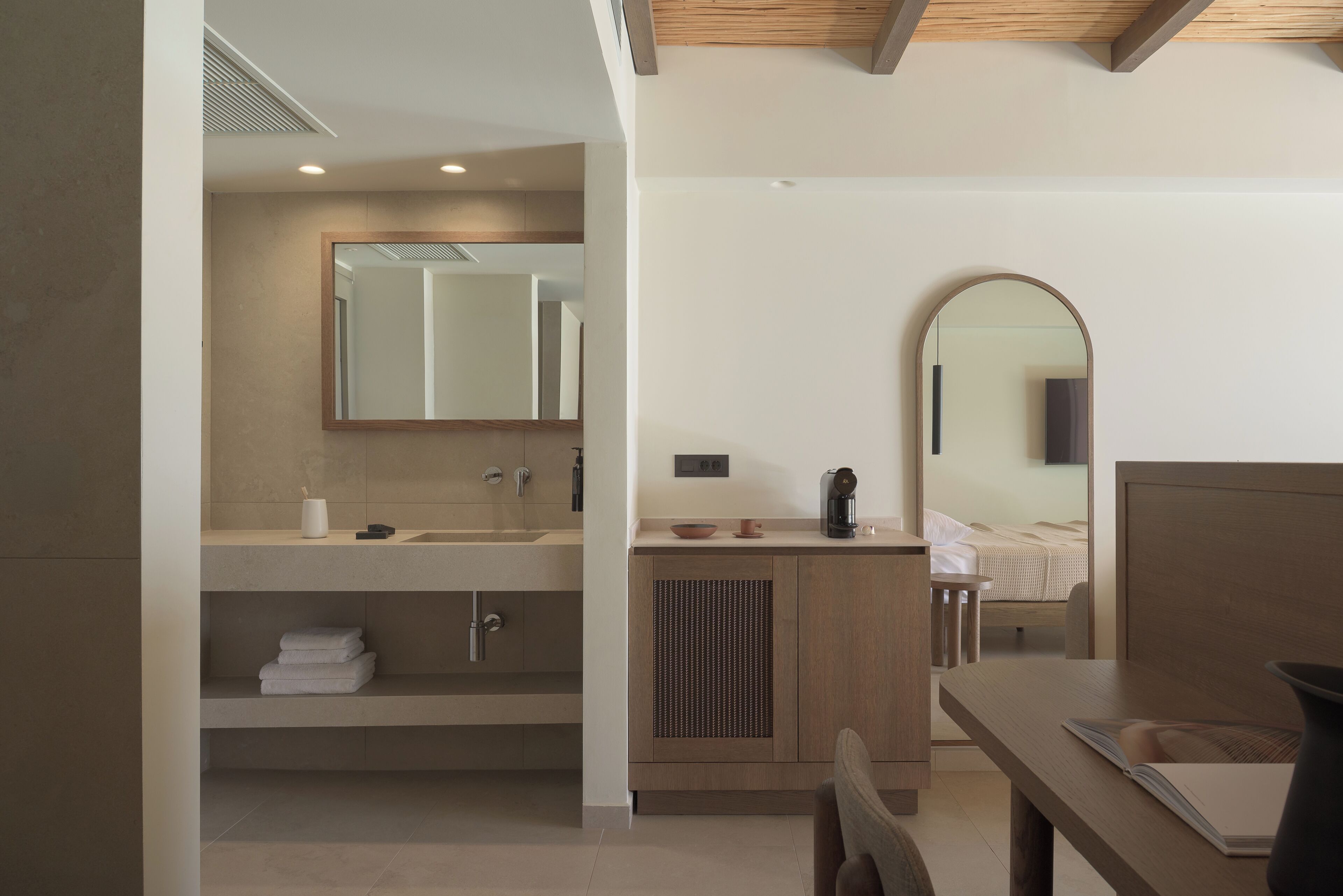 A modern bathroom with a large mirror, a sink, and a toilet. There is a wooden vanity and a bedside table with a book.
